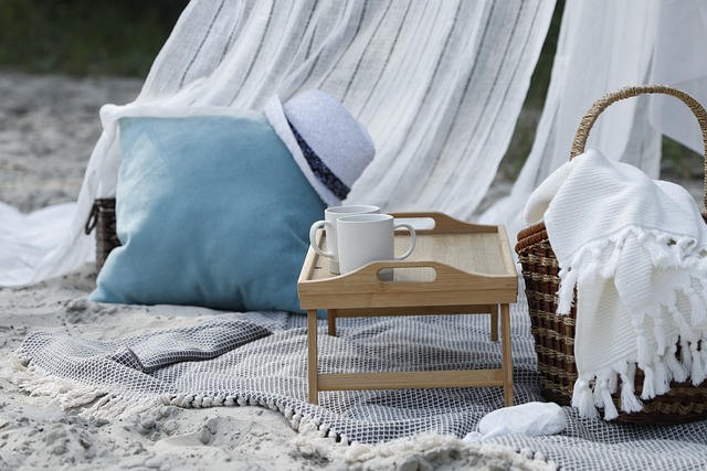 A soft white sandy beach scene with a wooden food tray and woven picnic basket atop a woven gray blanket. In the background are some gauzy white curtains, a blue pillow and a white hat.