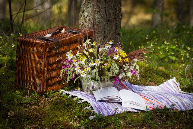 A blanket with a book and wild flowers sits in a grassy part of the woods next to a dark woven picnic basket.