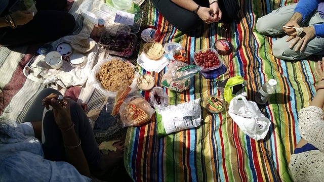 A bunch of people sit on a colorful striped picnic blanket. Only their hands and legs are pictured and an array of fun and delicious foods are laid out before them on the center of the blanket.