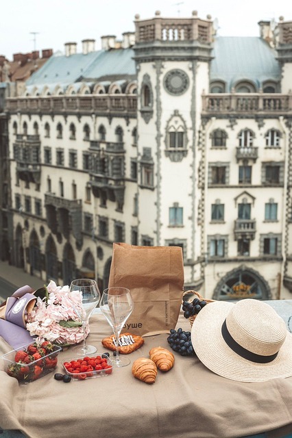 A brown blanket lies over a ledge looking out at a beautiful view of a European city. There are various fruits and foods as well as wine glasses arranged elegantly on the blanket.