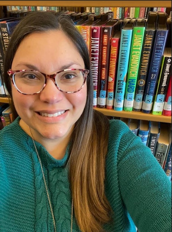 Victoria in a green sweater posed in front of Janet Evanovich's shelf in the mystery section.