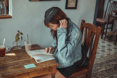 A woman in a denim jacket sits at a wooden table. She stares down at a book as she simultaneously turns the page and tucks her hair behind her ear. She appears to be in a cafe.  