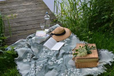 a green gray blanket sits next to the edge of a lake in the grass close to a wooden pier. There is a brown picnic basket, a straw hat, and a couple of books and a bottle of wine.