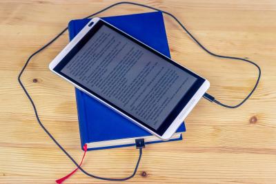 An eReader with text displayed on the screen is hooked up to a blue print book with a cord.